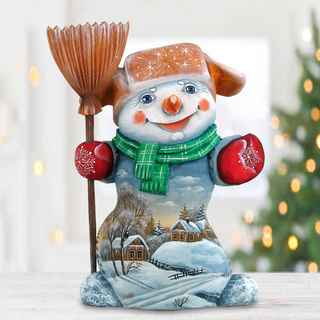 A snowman figurine with a green scarf and red gloves, holding a broom, is sitting on a snowy landscape with a house and trees in the background.