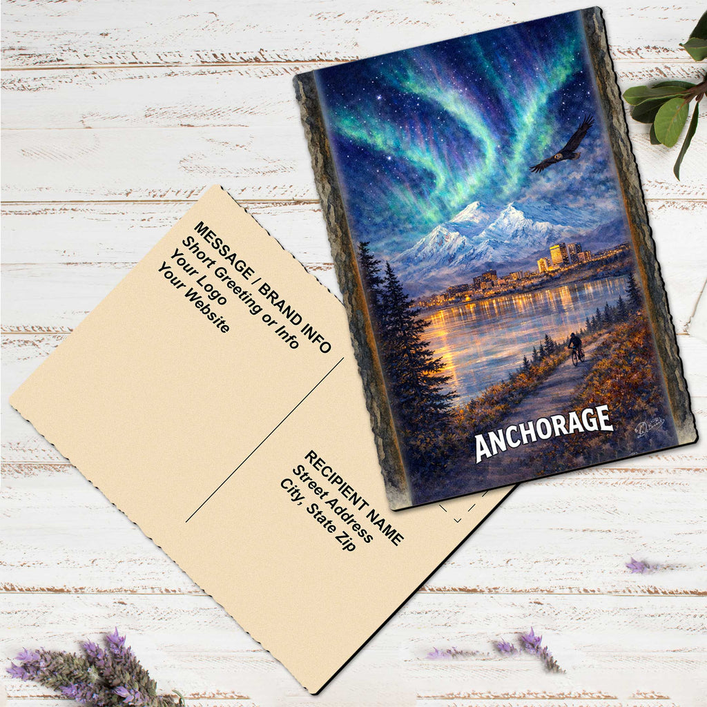 a postcard with a painting of a scenic landscape, including mountains, a lake, and a colorful sky with the Aurora Borealis. The postcard is placed on a white wooden surface, and there are some purple flowers nearby.