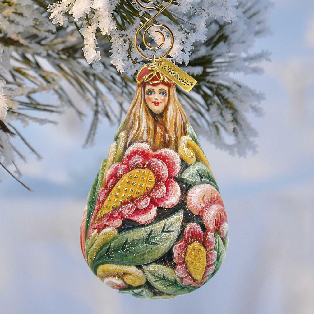 A colorful, hand-painted Christmas ornament with a woman's face and flowers hangs from a tree branch against a snowy background.