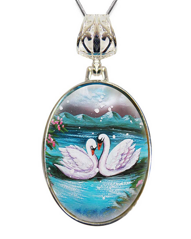 A pendant necklace with a circular oval-shaped design featuring two swans swimming in a body of water, surrounded by flowers and mountains in the background.