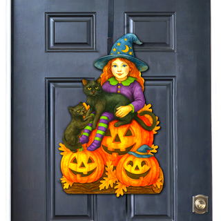 A Halloween-themed door decoration featuring a young girl in a witch's hat sitting on a pile of carved pumpkins and two black cats.