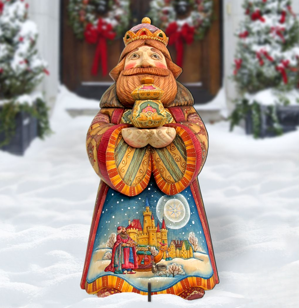 A colorful wooden statue of a king or queen, holding a crown and a book, stands in the snow in front of a wreath-adorned door.