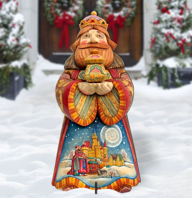 A colorful wooden statue of a king or queen, holding a crown and a book, stands in the snow in front of a wreath-adorned door.