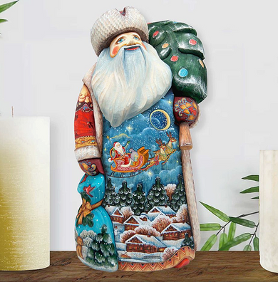 A colorful, hand-carved wooden Santa Claus figure with a white beard and hat, holding a green sack and a sled with a reindeer, set against a snowy winter landscape with a full moon and stars.