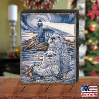 A wooden block with a painting of two otters swimming in the ocean, with a lighthouse in the background.