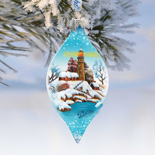 A blue glass ornament hanging from a tree branch, depicting a snowy winter landscape with a lighthouse in the distance.