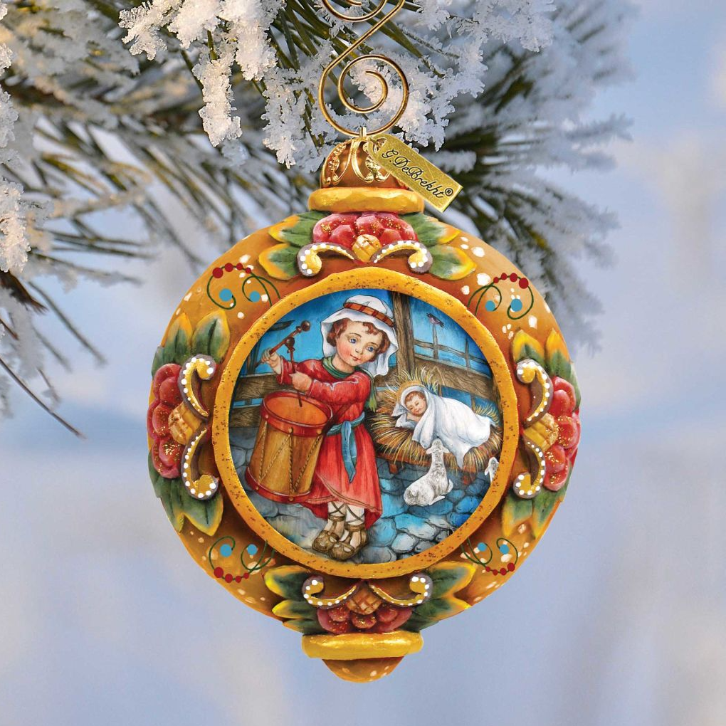 A decorative Christmas ornament featuring a young girl playing a drum, surrounded by a snowy landscape and a baby Jesus.