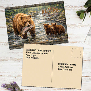 The image is a postcard featuring a painting of a bear catching a fish in a river, with two baby bears in the background. The postcard is placed on a white wooden surface, and there is a blank card below it with text for sending a message or providing information.
