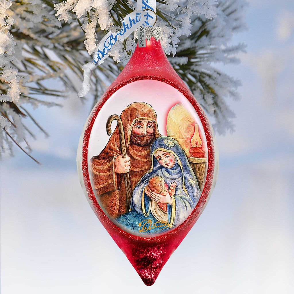 A red Christmas ornament with a nativity scene depicting the Virgin Mary and baby Jesus, hanging on a tree branch with snow.
