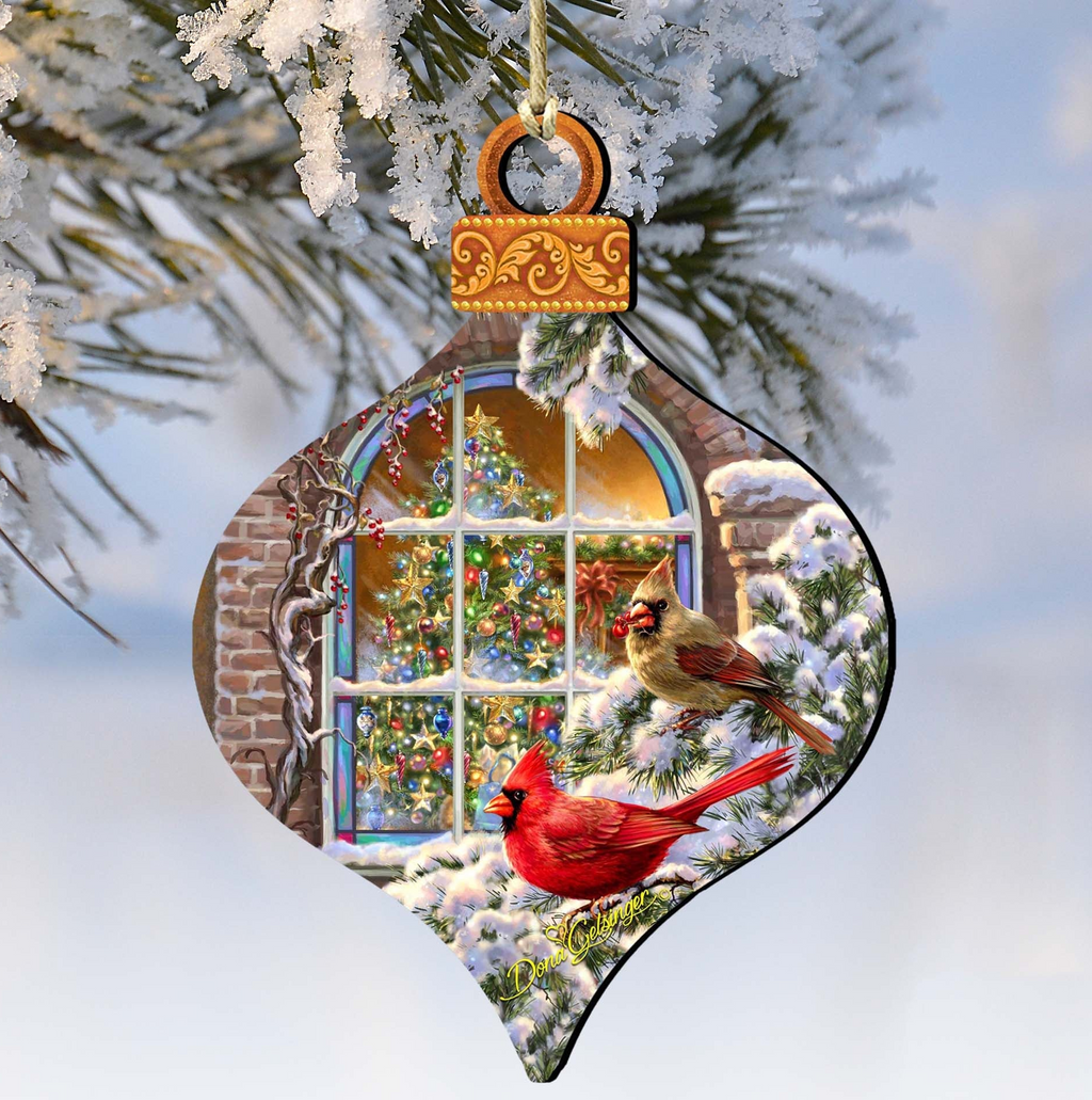 A decorative ornament featuring a snowy scene with two red cardinals perched on a branch, with a Christmas tree and a window visible through the branches.