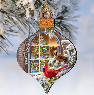 A decorative ornament featuring a snowy scene with two red cardinals perched on a branch, with a Christmas tree and a window visible through the branches.