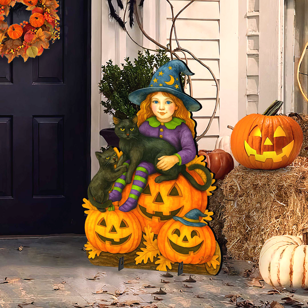 A Halloween-themed outdoor decoration featuring a young girl dressed as a witch, sitting on a pumpkin with a cat, and surrounded by jack-o-lanterns and hay bales.