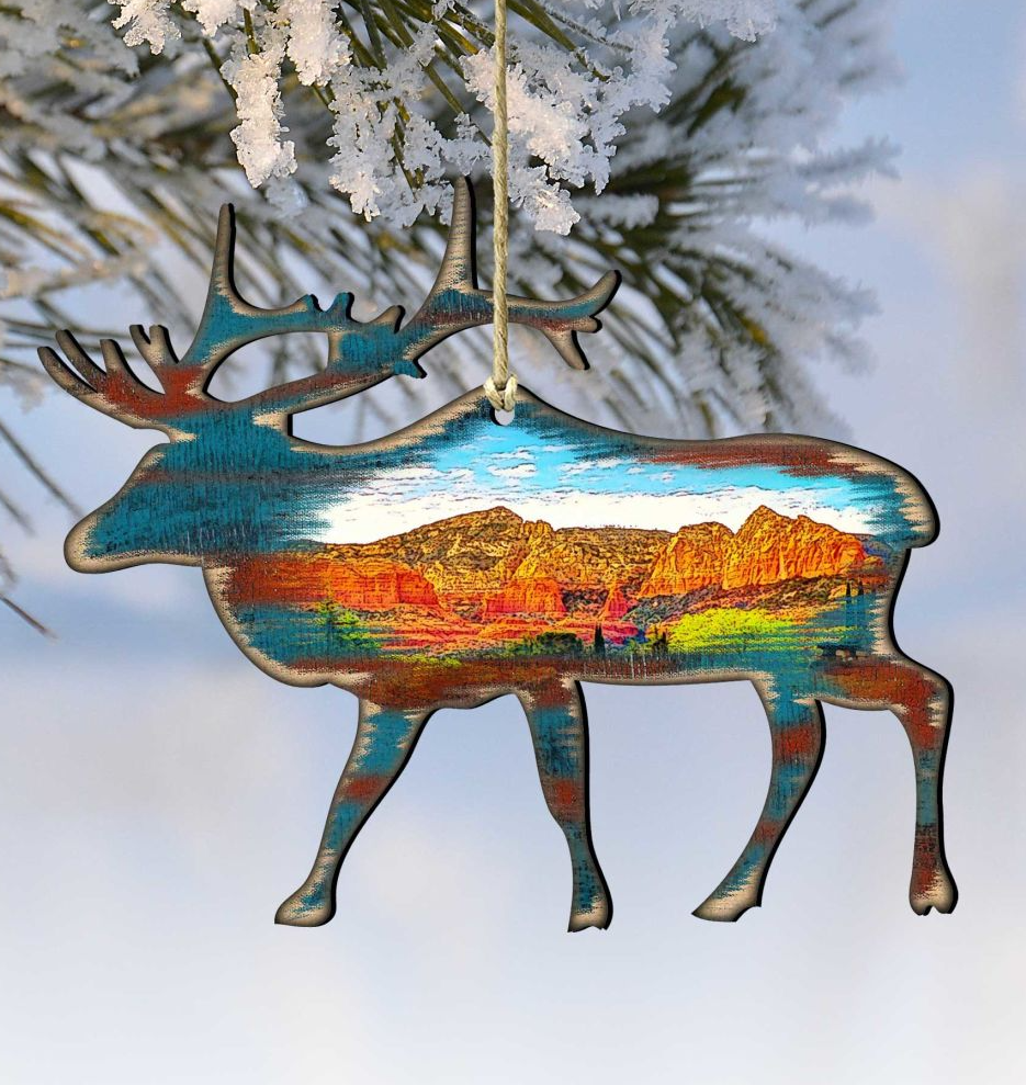 A wooden ornament in the shape of a deer, with a colorful landscape painted inside, hangs from a string against a snowy background.