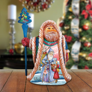 A figurine of Santa Claus holding a blue staff and a wreath, with a snowy landscape and other Christmas decorations in the background.