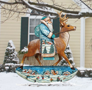 A wooden sculpture of Santa Claus riding a reindeer in a snowy landscape, with a house and trees in the background.