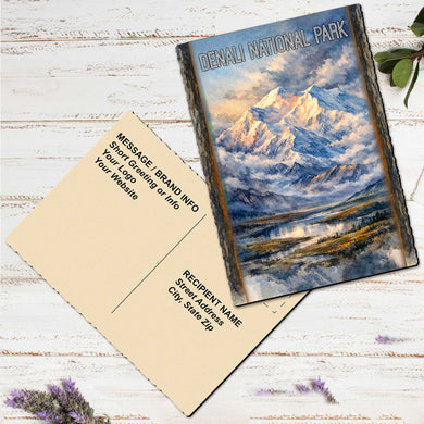 a postcard with a mountain landscape printed on it, placed on a white wooden surface alongside a blank postcard.