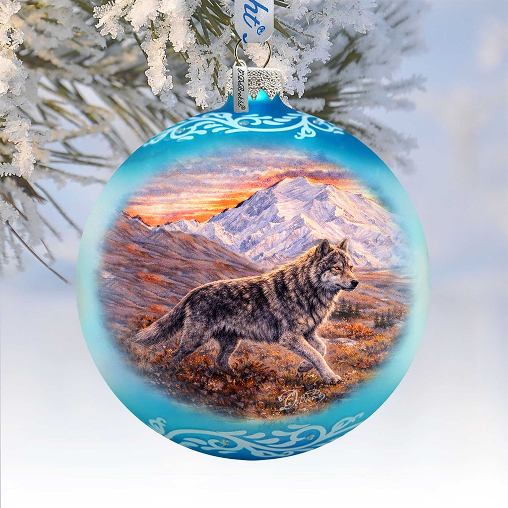 A blue ornament with a painting of a wolf walking through a snowy landscape, with mountains in the background.