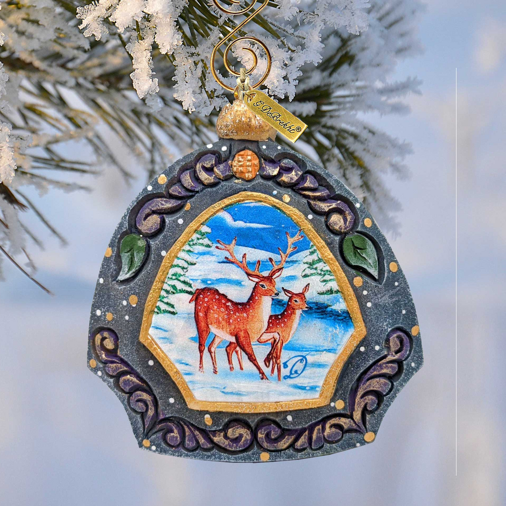 A decorative ornament featuring a deer standing in a snowy landscape, with a blue and gold frame and a gold-colored ornament hanging from a branch.