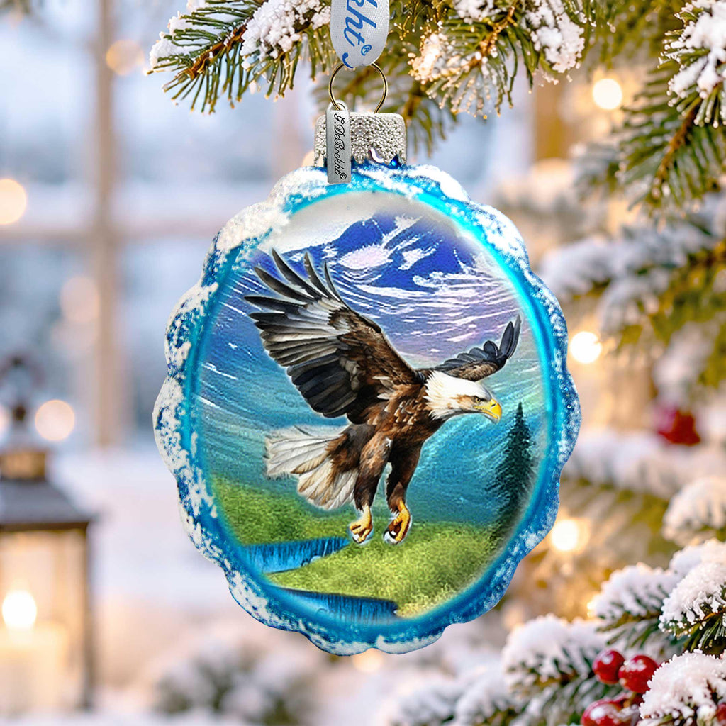 A blue ornament with a picture of a bald eagle in mid-flight, hanging on a snowy branch of a Christmas tree.