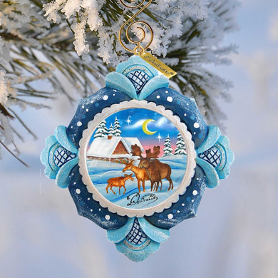 A decorative ornament hanging from a tree branch, featuring a snowy winter scene with a cabin, reindeer, and a crescent moon.