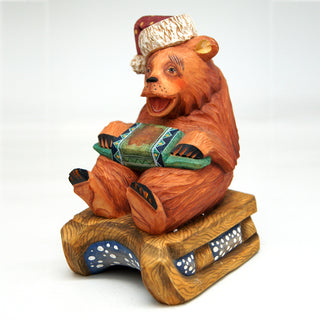 A wooden bear figurine wearing a hat and holding a musical instrument, sitting on a wooden sled.