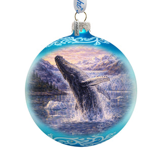 A blue glass ornament with a painting of a whale jumping out of the water, surrounded by mountains and a sunset sky.