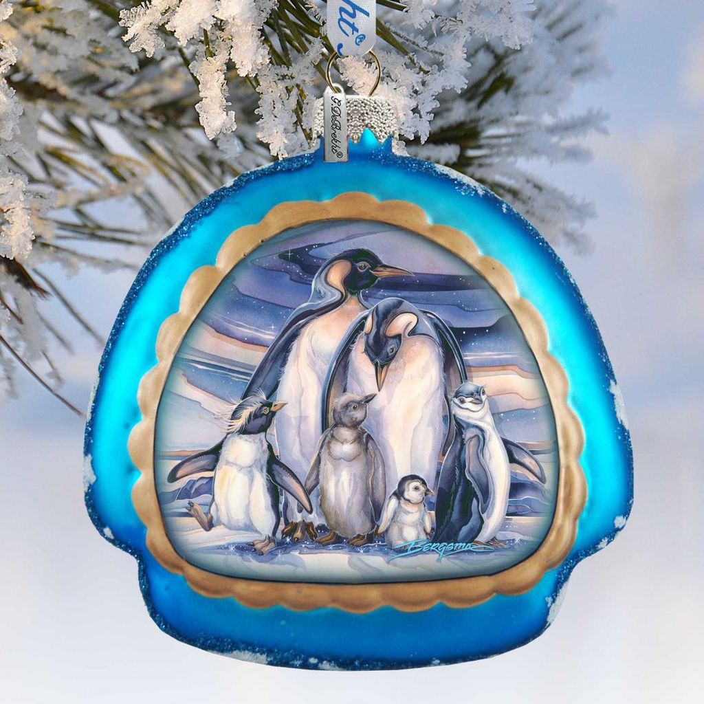 A blue ornament with a picture of a group of penguins on it, hanging on a tree branch covered in snow.