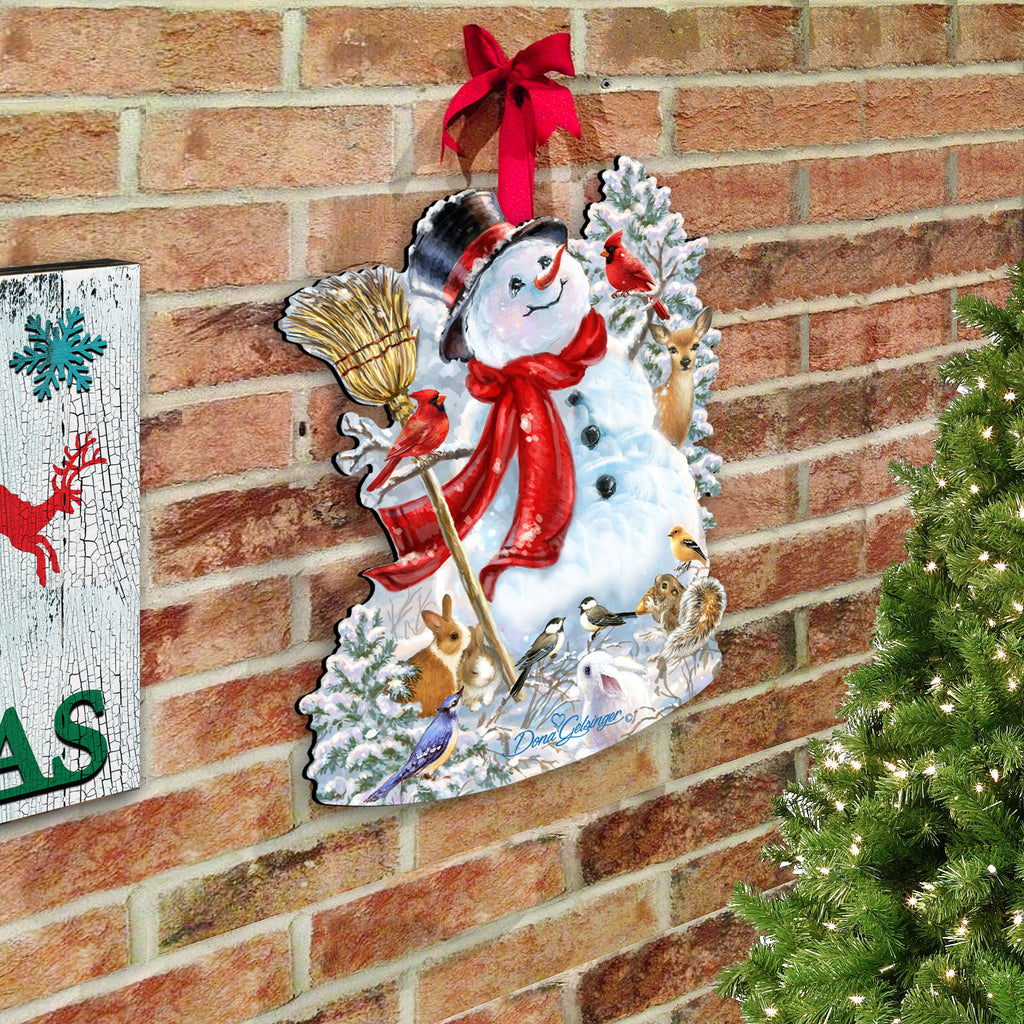 A festive Christmas scene with a snowman, a red bow, and various holiday decorations such as a bird, a rabbit, and a reindeer. The snowman is surrounded by a wreath of snow and pine branches, and the scene is set against a brick wall.