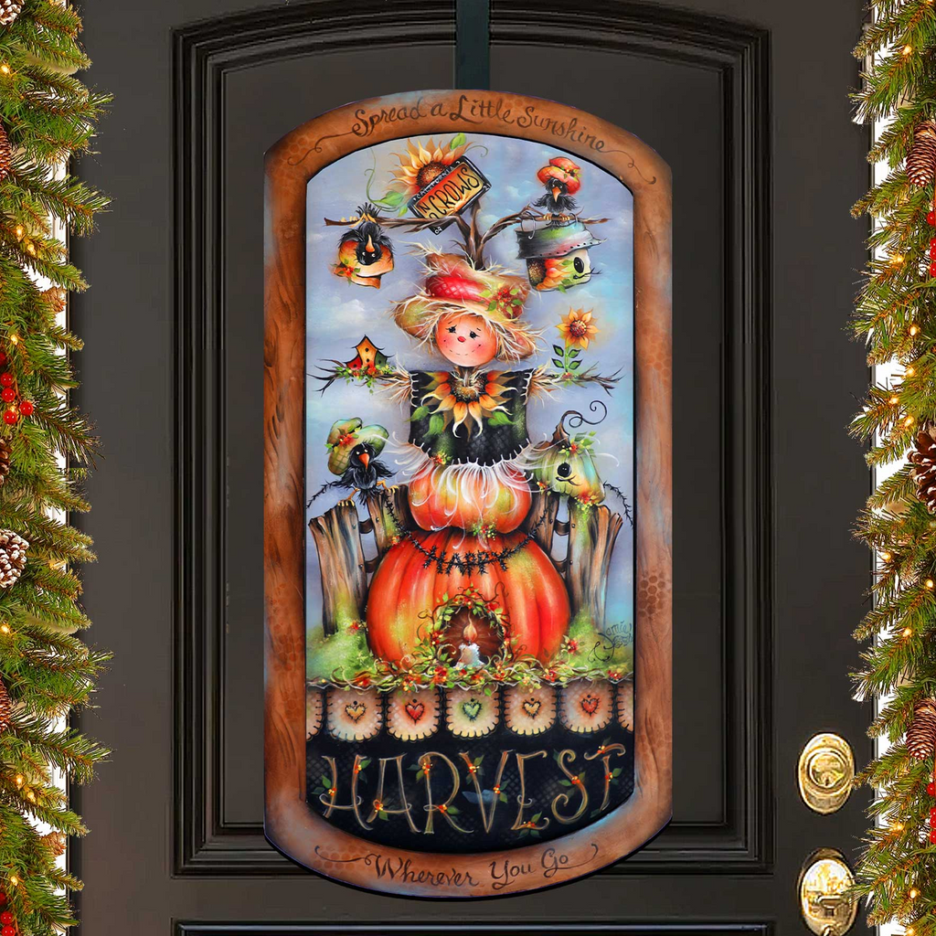a decorative sign hanging on a door, depicting a scarecrow with a pumpkin and other fall-themed elements.