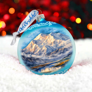 A blue Christmas ornament with a painting of a mountain landscape, featuring a snowy mountain and a body of water, is placed on a snowy surface against a background of red and yellow lights.