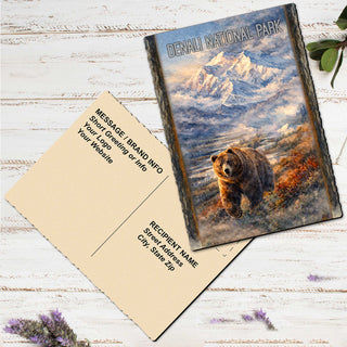 a postcard with a painting of a bear in a mountainous landscape, placed on a white wooden surface alongside a blank postcard.