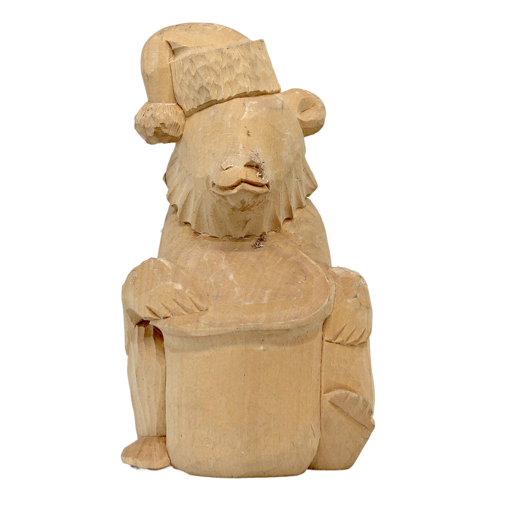 A wooden carving of a bear wearing a hat and holding a hat in its paws, sitting on a hat.