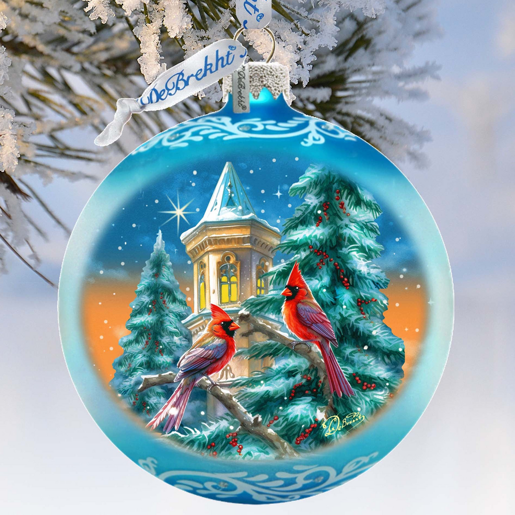 A blue Christmas ornament with a snowy scene featuring two red cardinals perched on a branch, with a building and starry sky in the background.