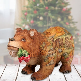 A wooden bear figurine with a painted design of bears and berries, placed on a wooden surface in front of a blurred Christmas tree.