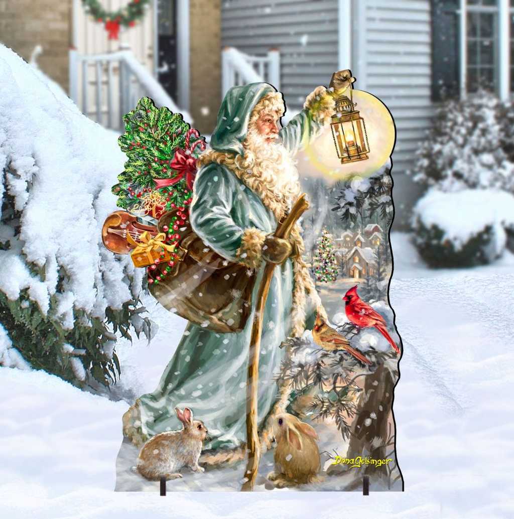 A cardboard cutout of Santa Claus holding a lantern and a bag of presents, surrounded by snow and birds, with a wreath and a house in the background.
