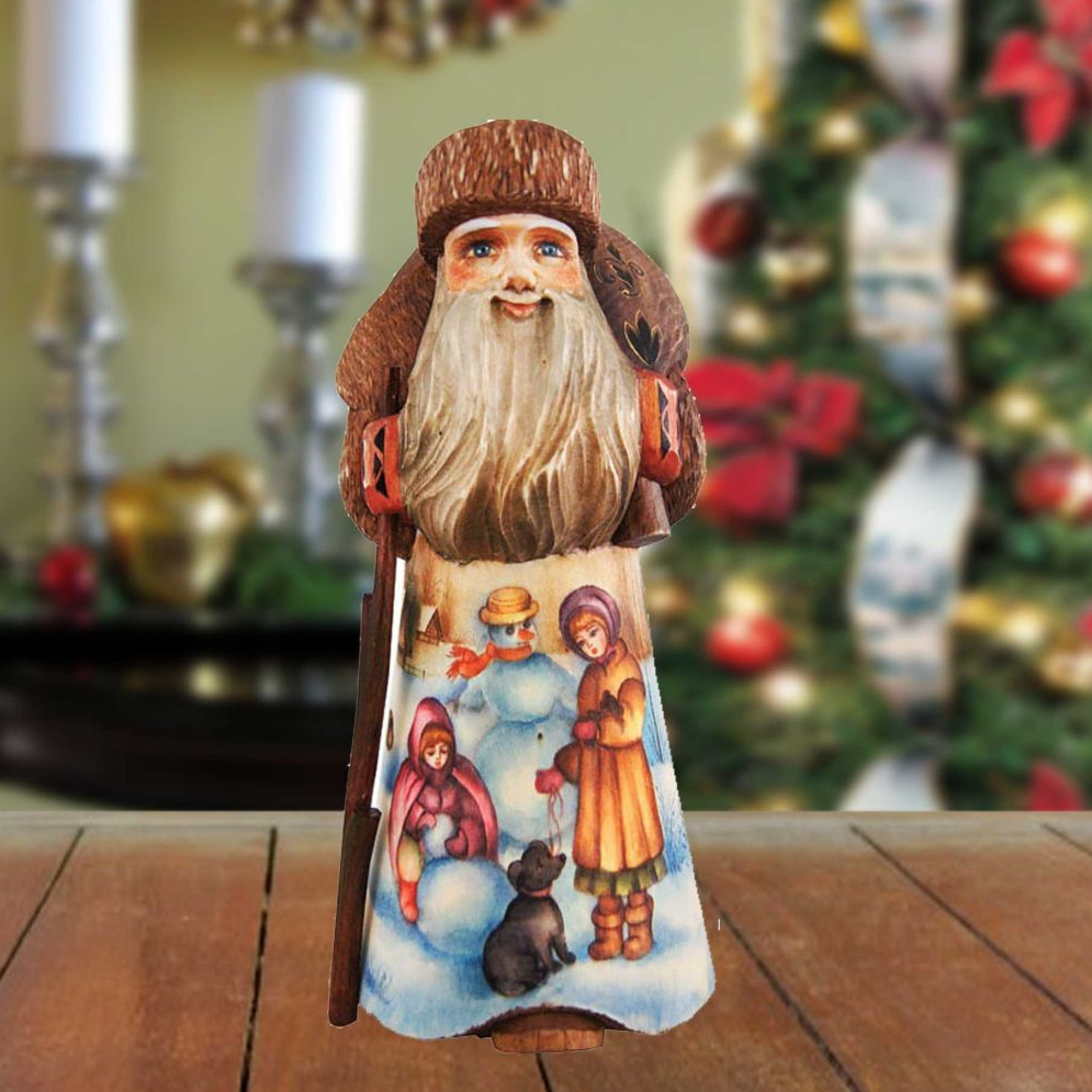 A wooden Santa Claus figurine with a long beard and hat, standing on a wooden surface with a Christmas tree and decorations in the background.