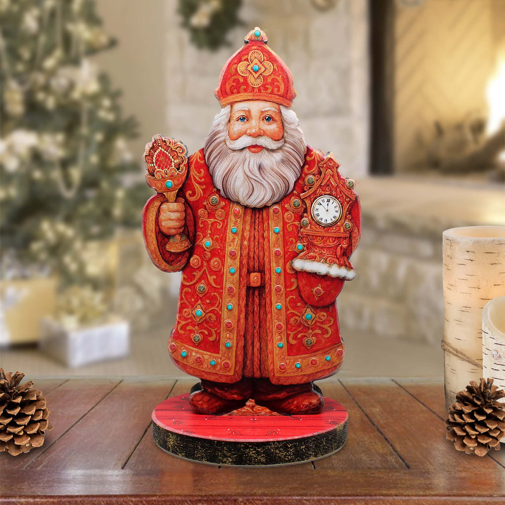 A figurine of Santa Claus wearing a red and gold robe and hat, holding a clock, stands on a wooden surface with pine cones and a blurred background.