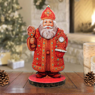 A figurine of Santa Claus wearing a red and gold robe and hat, holding a clock, stands on a wooden surface with pine cones and a blurred background.