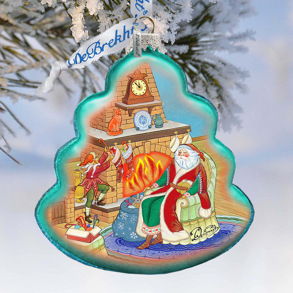A colorful ornament depicting Santa Claus sitting in a chair by a fireplace, with a clock on the mantel and a small cat on the mantel.