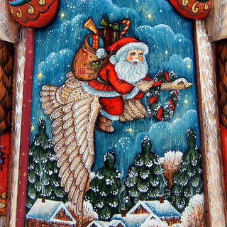 A colorful painting depicting Santa Claus riding a large bird, carrying a sack of presents, flying over a snowy landscape with trees and houses below.