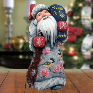 A wooden Santa Claus figurine holding a snowflake ornament and a bird ornament stands on a wooden surface, with a blurred Christmas tree and decorations in the background.