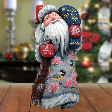 A wooden Santa Claus figurine holding a snowflake ornament and a bird ornament stands on a wooden surface, with a blurred Christmas tree and decorations in the background.