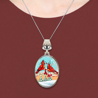 A silver necklace with a circular pendant featuring a painting of two red cardinals perched on a snowy branch.