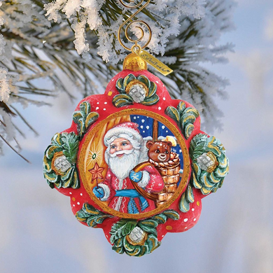 A colorful ornament featuring a smiling Santa Claus figure, a teddy bear, and a star, hanging from a tree branch against a snowy background.