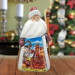 A wooden Santa Claus figurine holding a staff and a decorated nativity scene, with a blurred Christmas tree in the background.