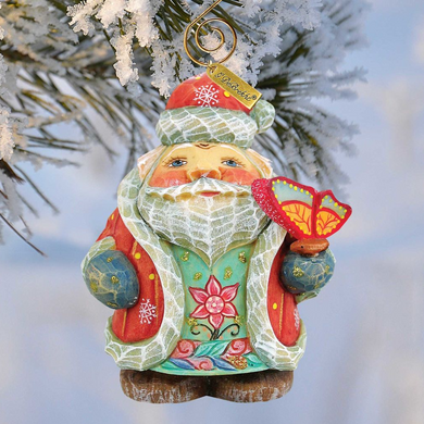 A colorful ornament of a smiling Santa Claus figure holding a butterfly, hanging from a tree branch covered in snow.