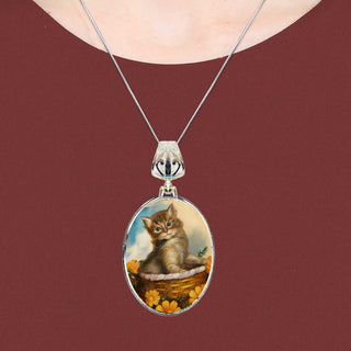 A silver necklace with a pendant featuring a painting of a kitten sitting in a basket.